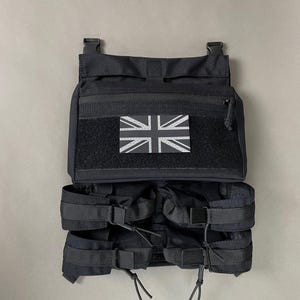 Könnte beinhalten: Schwarze taktische Weste mit mehreren Taschen und verstellbaren Riemen. Verfügt über einen Klettverschluss mit einer Union Jack-Flagge. Die Weste hat eine Reißverschlusstasche und ist für das Tragen von Ausrüstung konzipiert.