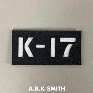 Könnte beinhalten: Schwarzer rechteckiger Aufnäher mit dem weißen Text "K-17" und dem Text "A.R.K SMITH" unten. Die Zahlen und Buchstaben sind ausgeschnitten und zeigen ein Wabenmuster.