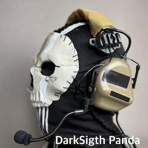 Puede incluir: Un pasamontañas negro con una máscara de calavera y auriculares. La máscara de calavera es blanquecina con costuras negras y una franja blanca. Los auriculares son de color canela con un micrófono negro. El texto "DarkSigth Panda" es visible.