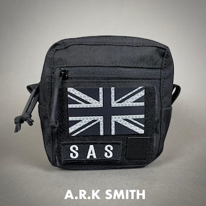 May include: Black tactical pouch with a Union Jack patch featuring a reflective honeycomb pattern. The pouch has a zippered main compartment and a smaller patch with the letters "SAS". The text "A.R.K SMITH" is printed at the bottom.
