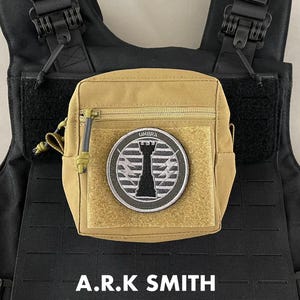 Könnte beinhalten: Eine beige taktische Tasche mit Reißverschluss und Klettverschluss. Der Aufnäher zeigt ein schwarz-weißes Design mit dem Wort "UMBRA" über einem stilisierten Turm. Die Tasche ist an einer schwarzen taktischen Weste befestigt. Der Text "A.R.K SMITH" steht unten.
