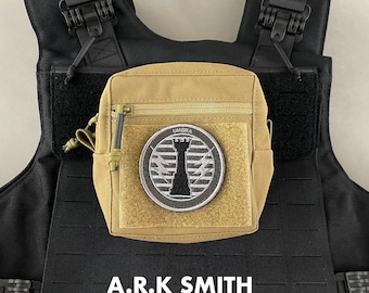 May include: A tan tactical pouch with a zipper closure and a velcro patch. The patch features a black and white design with the word "UMBRA" above a stylized tower. The pouch is attached to a black tactical vest. The text "A.R.K SMITH" is at the bottom.