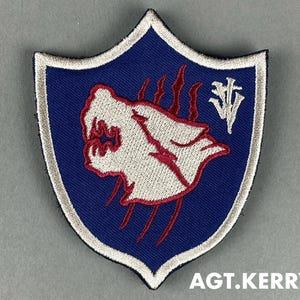 Puede incluir: Parche bordado con forma de escudo, con una criatura blanca con forma de lobo y detalles rojos sobre un fondo azul. El parche tiene un borde blanco y el texto "AGT.KERRY" en la parte inferior.