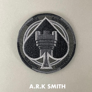 以下が含まれることがあります： 黒いスペードのエースのデザインと中央に城の塔がある、丸い刺繍パッチ。パッチはグレーとオリーブグリーンの配色で、下部に「A.R.K SMITH」の文字があります。