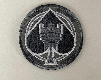 May include: A round, embroidered patch featuring a black ace of spades design with a castle tower in the center. The patch has a gray and olive green color scheme, with the text "A.R.K SMITH" at the bottom.
