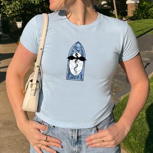 May include: Pale blue crew neck t-shirt featuring a stained glass dragon design. The graphic shows a black dragon, a white egg, and a blue and white stained glass frame. The shirt is worn with light wash blue jeans.