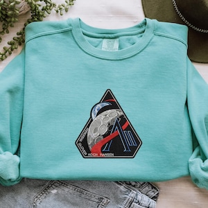 May include: A light teal crew neck sweatshirt with a black, red, and white patch depicting a moon and planet. The patch includes the names: Wiseman, Glover, Koch, and Hansen. The sweatshirt is folded on top of a pair of grey jeans.