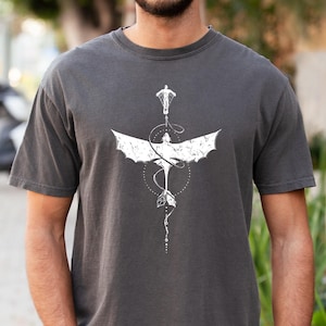 May include: Dark gray t-shirt featuring a white graphic of a stylized dragon with outstretched wings and a figure at the top. The design includes a dotted line and geometric shapes. The shirt has short sleeves.