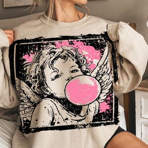 May include: Beige sweatshirt featuring a black and pink graphic of an angel blowing a bubblegum bubble. The angel has wings and is set against a pink splatter background within a black rectangular frame. The sweatshirt is a neutral color.