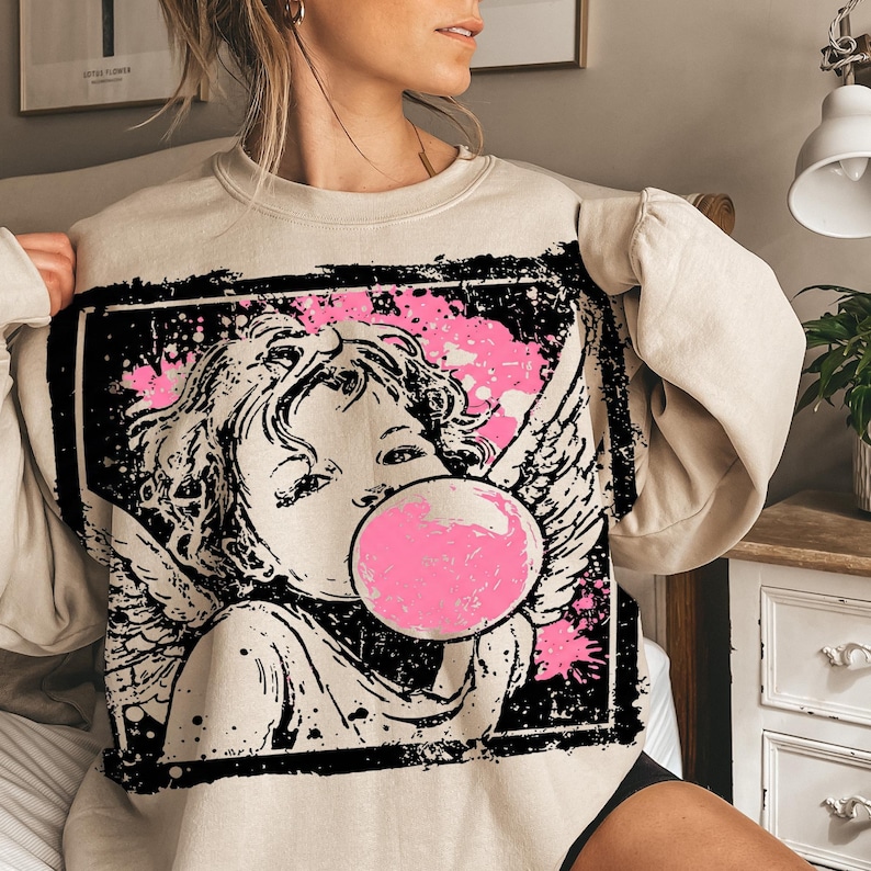 May include: Beige sweatshirt featuring a black and pink graphic of an angel blowing a bubblegum bubble. The angel has wings and is set against a pink splatter background within a black rectangular frame. The sweatshirt is a neutral color.