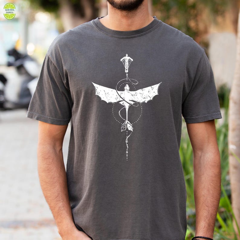 May include: Dark grey t-shirt with a white dragon graphic. The design includes a figure, wings, and geometric shapes. The shirt has short sleeves. The dragon graphic is centrally placed on the chest.