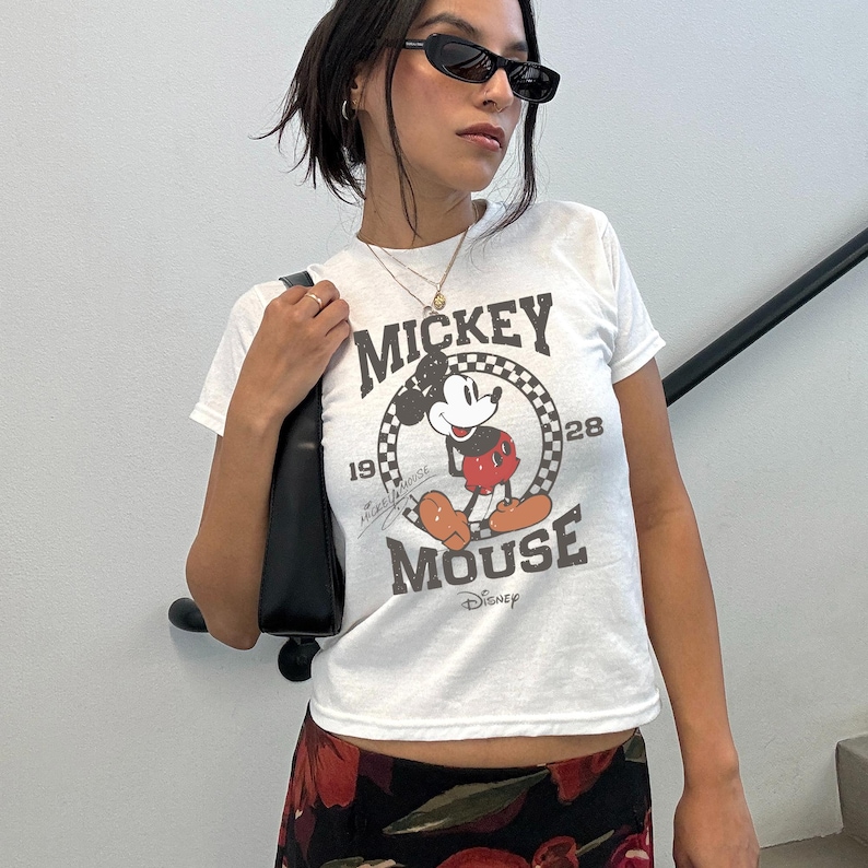 May include: White t-shirt with a Mickey Mouse graphic. The design includes the words "Mickey Mouse" and the number "28" within a chequered circle. The shirt has short sleeves.