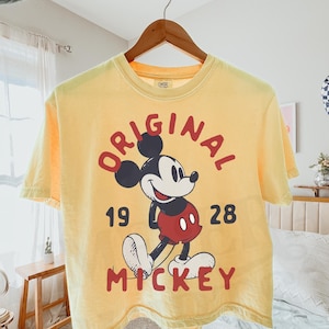 May include: Yellow t-shirt featuring a graphic of Mickey Mouse with the word "ORIGINAL" above the character and the words "19 28 MICKEY" below. The shirt is hanging on a wooden hanger.