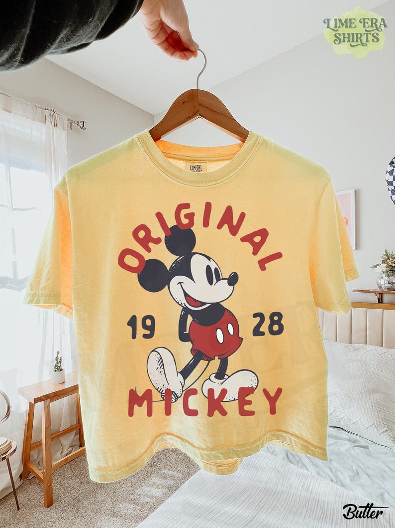 May include: Yellow t-shirt featuring a graphic of Mickey Mouse with the word "ORIGINAL" above the character and the words "19 28 MICKEY" below. The shirt is hanging on a wooden hanger.