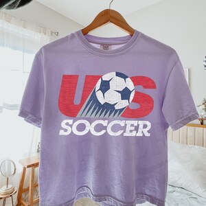 May include: A light purple short-sleeve t-shirt featuring a graphic with the red text US and the word SOCCER in white below it. A black and white soccer ball graphic is positioned over the letter S with motion lines trailing behind it.