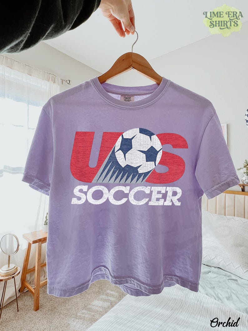 May include: A light purple short-sleeve t-shirt featuring a graphic with the red text US and the word SOCCER in white below it. A black and white soccer ball graphic is positioned over the letter S with motion lines trailing behind it.