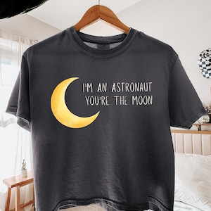 May include: A charcoal grey short-sleeve t-shirt featuring a yellow crescent moon graphic on the left side. To the right of the moon, the white text reads: I'M AN ASTRONAUT YOU'RE THE MOON. The shirt is displayed on a wooden hanger against a neutral background.