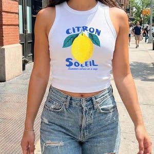 Puede incluir: Una camiseta de tirantes blanca acanalada con un gr&aacute;fico de un lim&oacute;n amarillo con hojas verdes. El texto sobre el lim&oacute;n dice CITRON en azul, y el texto debajo dice SOLEIL en azul, seguido del texto m&aacute;s peque&ntilde;o Summer citrus in a cup.