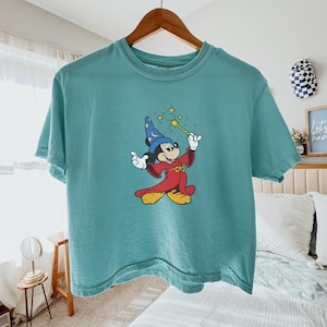 May include: Teal-colored t-shirt featuring a graphic of Mickey Mouse dressed as a wizard, holding a wand with stars. Mickey is wearing a blue wizard hat and a red robe. The shirt is hanging on a wooden hanger.