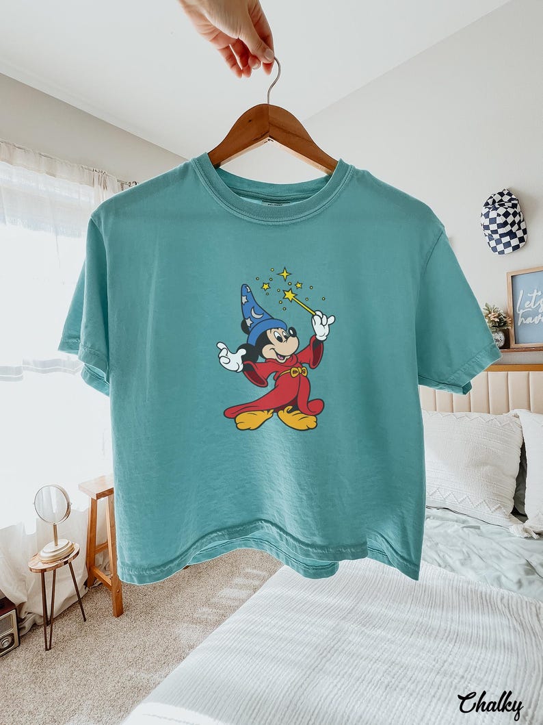 May include: Teal-colored t-shirt featuring a graphic of Mickey Mouse dressed as a wizard, holding a wand with stars. Mickey is wearing a blue wizard hat and a red robe. The shirt is hanging on a wooden hanger.