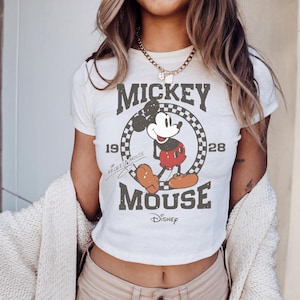 May include: Cream-coloured crop top with a distressed Mickey Mouse graphic. The design includes the text "Mickey Mouse" and "1928" with a chequered circle. The Disney logo is at the bottom.