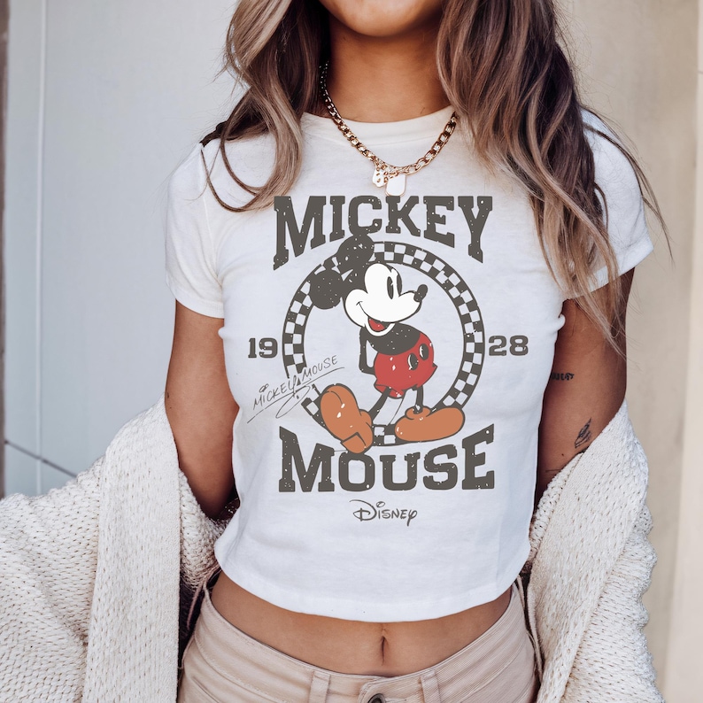May include: Cream-coloured crop top with a distressed Mickey Mouse graphic. The design includes the text "Mickey Mouse" and "1928" with a chequered circle. The Disney logo is at the bottom.