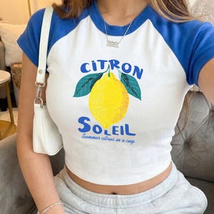 Puede incluir: Una camiseta ragl&aacute;n blanca y azul con un gr&aacute;fico de un lim&oacute;n amarillo con hojas verdes. El texto "CITRON SOLEIL" aparece en letras azules sobre y bajo el lim&oacute;n, con "Summer citrus in a cup" escrito en letra peque&ntilde;a azul debajo.