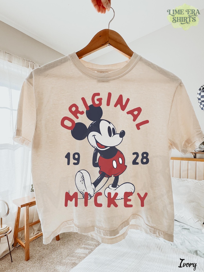 May include: Ivory-colored t-shirt featuring a graphic of Mickey Mouse with the words "ORIGINAL MICKEY 1928" in red. The shirt is hanging on a wooden hanger. The brand name "Lime Era Shirts" is visible in the top right corner.