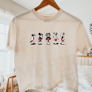 Puede incluir: Una camiseta de manga corta de color marfil con un estampado gr&aacute;fico de cinco figuras de Mickey Mouse en varias poses de baile, cada una con sombrero y chaqueta. La camiseta se muestra en una percha de madera sobre un fondo claro.
