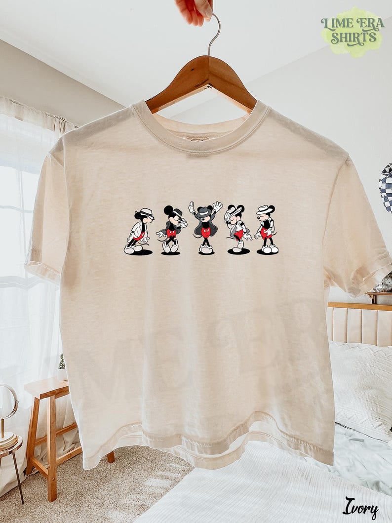Puede incluir: Una camiseta de manga corta de color marfil con un estampado gr&aacute;fico de cinco figuras de Mickey Mouse en varias poses de baile, cada una con sombrero y chaqueta. La camiseta se muestra en una percha de madera sobre un fondo claro.