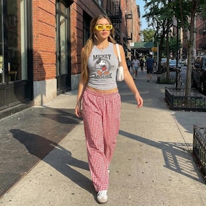 May include: A grey vest top featuring a Mickey Mouse design, worn with red and white gingham trousers. The outfit is accessorized with yellow sunglasses and white trainers. The person is walking on a pavement in a city.