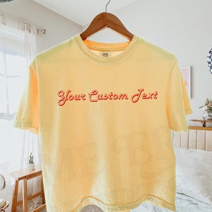 May include: A pale yellow short-sleeve t-shirt featuring the red text Your Custom Text printed in a retro cursive font across the chest. The shirt is displayed on a wooden hanger against a light-colored interior setting with a visible brand tag.