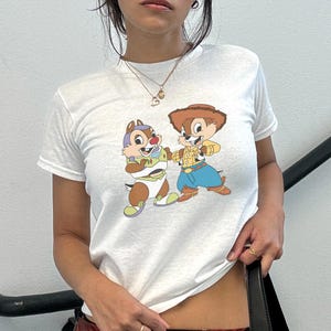 May include: White t-shirt featuring a cartoon graphic of two chipmunks dressed as characters from Toy Story. One chipmunk is in a Buzz Lightyear costume, and the other is dressed as Woody. The shirt has a crew neck and short sleeves.