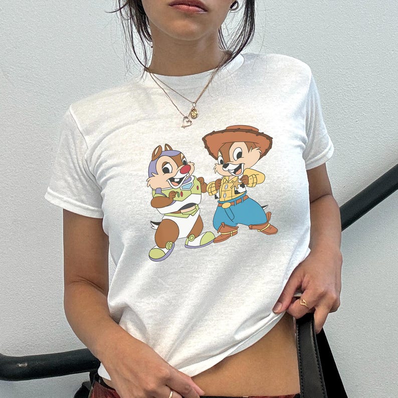 May include: White t-shirt featuring a cartoon graphic of two chipmunks dressed as characters from Toy Story. One chipmunk is in a Buzz Lightyear costume, and the other is dressed as Woody. The shirt has a crew neck and short sleeves.