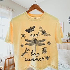 May include: A pale yellow short-sleeve t-shirt featuring a black ink-style graphic of various insects including a dragonfly, moth, and beetle. The text "sad bug summer" is printed in a cursive font in the centre among the insect illustrations.
