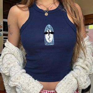 May include: Navy blue ribbed tank top featuring a dragon design within a stained glass window. The top has a round neck and is sleeveless. The dragon is black with a white background and blue detailing.