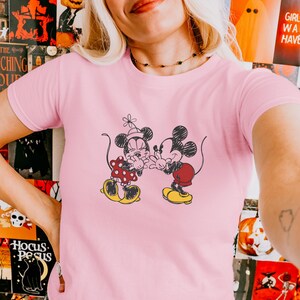 May include: Pink t-shirt featuring a black and white illustration of Mickey and Minnie Mouse. Minnie wears a red polka dot dress and Mickey wears red shorts. Both characters are in a loving embrace, with yellow shoes.
