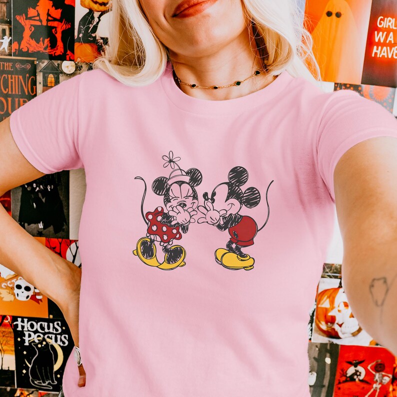 May include: Pink t-shirt featuring a black and white illustration of Mickey and Minnie Mouse. Minnie wears a red polka dot dress and Mickey wears red shorts. Both characters are in a loving embrace, with yellow shoes.