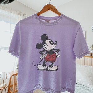 以下が含まれることがあります： ミッキーマウスのグラフィックが特徴のラベンダー色のTシャツ。漫画のキャラクターは黒、白、赤で描かれています。Tシャツは木製のハンガーに掛けられています。「Orchid」の文字が右下に印刷されています。