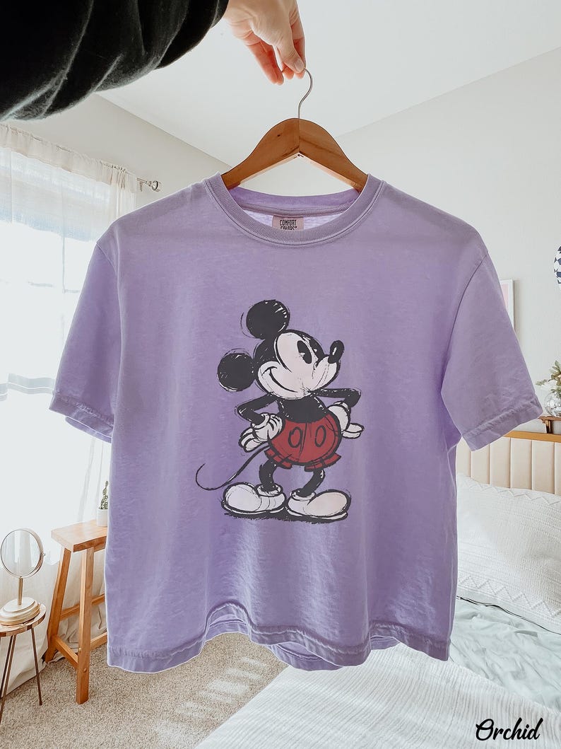 以下が含まれることがあります： ミッキーマウスのグラフィックが特徴のラベンダー色のTシャツ。漫画のキャラクターは黒、白、赤で描かれています。Tシャツは木製のハンガーに掛けられています。「Orchid」の文字が右下に印刷されています。