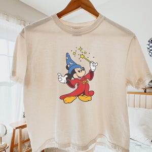 May include: Ivory-colored t-shirt featuring a graphic of Mickey Mouse as a sorcerer, holding a wand with stars. Mickey is wearing a blue hat, red robe, and yellow shoes. The shirt is hanging on a wooden hanger.
