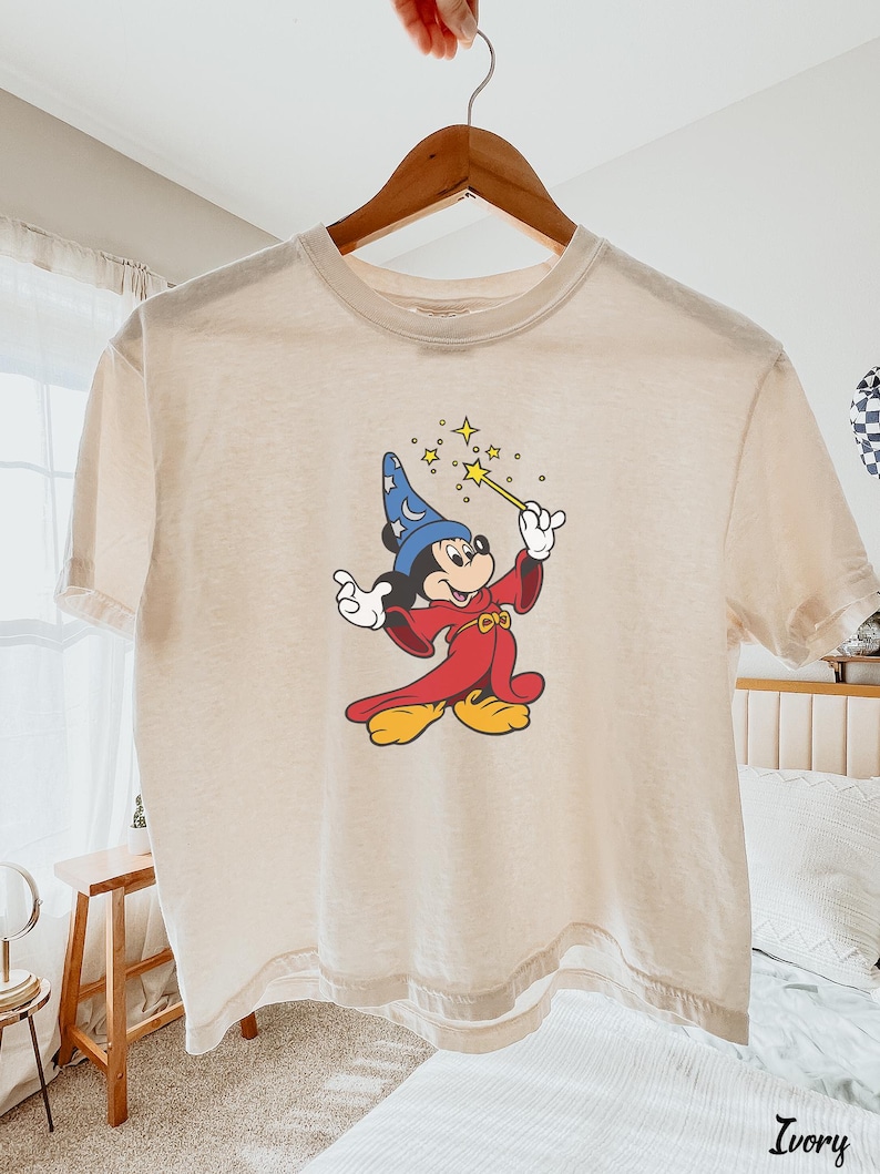 May include: Ivory-colored t-shirt featuring a graphic of Mickey Mouse as a sorcerer, holding a wand with stars. Mickey is wearing a blue hat, red robe, and yellow shoes. The shirt is hanging on a wooden hanger.