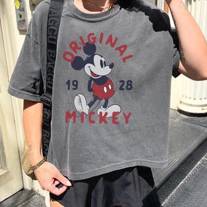 May include: Dark gray t-shirt featuring a classic Mickey Mouse graphic. The design includes the words "ORIGINAL MICKEY" and the year "1928" in red and navy blue. The shirt has a vintage, worn look.