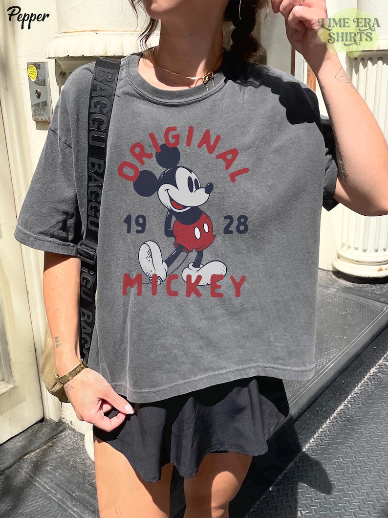 May include: Dark gray t-shirt featuring a classic Mickey Mouse graphic. The design includes the words "ORIGINAL MICKEY" and the year "1928" in red and navy blue. The shirt has a vintage, worn look.