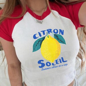 Puede incluir: Una camiseta blanca de manga corta ragl&aacute;n con mangas y cuello rojos. El frente presenta un gr&aacute;fico de un lim&oacute;n amarillo con hojas verdes, el texto azul "CITRON SOLEIL" y el texto azul m&aacute;s peque&ntilde;o "Summer citrus in a cup."