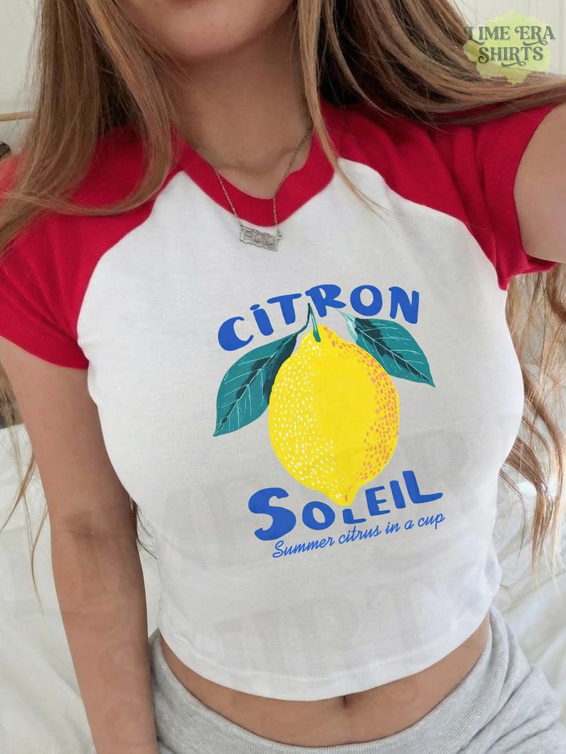 Puede incluir: Una camiseta blanca de manga corta ragl&aacute;n con mangas y cuello rojos. El frente presenta un gr&aacute;fico de un lim&oacute;n amarillo con hojas verdes, el texto azul "CITRON SOLEIL" y el texto azul m&aacute;s peque&ntilde;o "Summer citrus in a cup."
