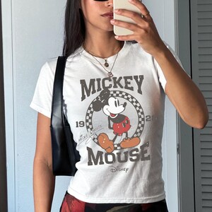 May include: White t-shirt with a distressed Mickey Mouse graphic. The design includes "Mickey Mouse" and "Disney" with a checkered circle around Mickey. The shirt has short sleeves.