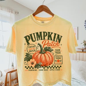 May include: A pale yellow t-shirt featuring a graphic print of a large orange pumpkin with green leaves and text that reads PUMPKIN Patch, FRESH FROM THE PATCH, FARM FRESH, LOCALLY GROWN, AUTUMN PICKED, PUMPKINS, CORN MAZE, and APPLE CIDER.