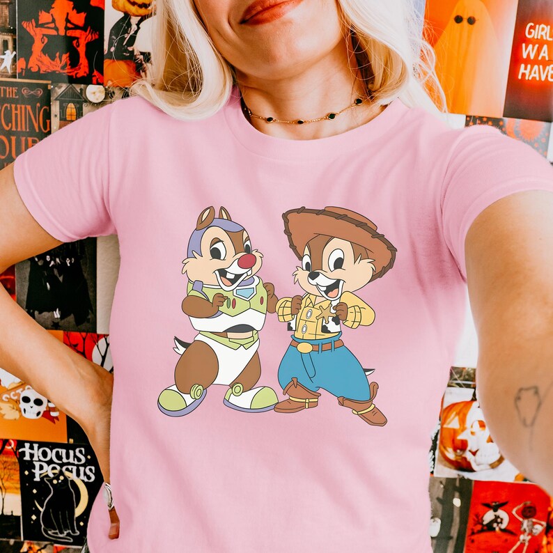 May include: Pink t-shirt featuring a cartoon graphic of two chipmunks dressed as Buzz Lightyear and Woody. The chipmunks are brown, tan, and white with blue, green, yellow, and red accents. The shirt is made of a soft material.