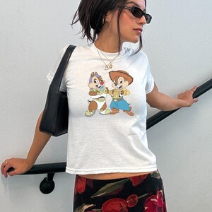 May include: White t-shirt featuring a cartoon graphic of two chipmunks. One chipmunk is dressed as Buzz Lightyear, and the other is dressed as Woody. The shirt has short sleeves and a crew neck.